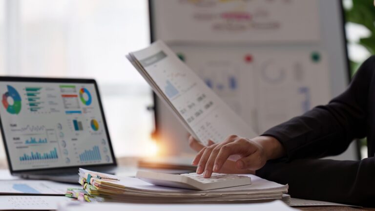 Woman sitting at a desk, reviewing accounting reports with a calculator and a laptop, analyzing financial data and preparing financial statements.