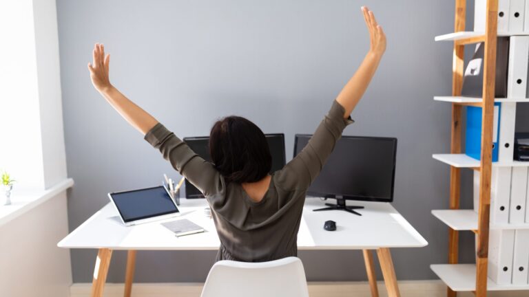 Woman working remotely with arms raised in celebration while seated at a desk with a laptop, viewed from behind.